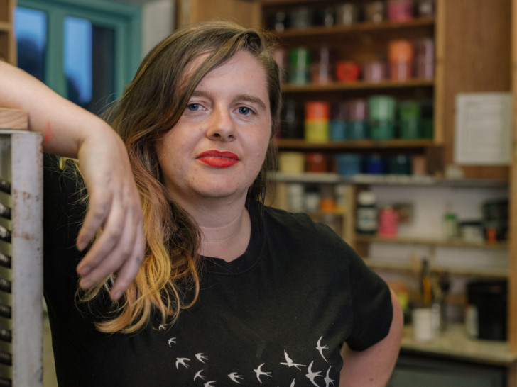 Photograph of a woman standing in an art studio, with long hair wearing a black t shirt and red lipstick