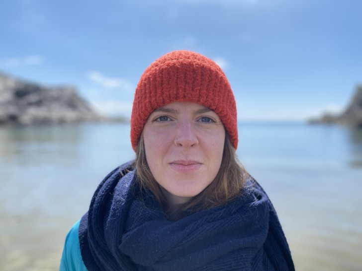 A woman wears a bright red hat and a navy scarf and is standing in front of a blue sea and sky with cliffs on either side.