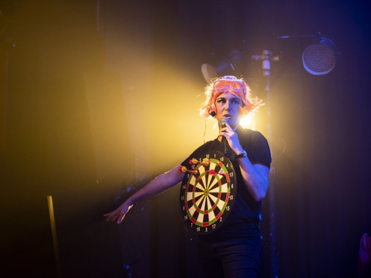 Amy Pennington is dressed in black and is wearing a bright pink wig. They have a dart board hung around their neck so that the target rests on their chest. They are on stage performing, holding a microphone.