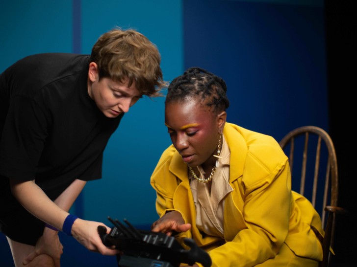 Nandi Bhebhe & Phoebe Davies are in a room with dark blue walls, they are photographed looking down at a device that Phoebe is holding.