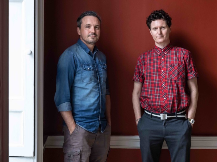 Two men standing in front of a red painted wall, they are looking at the camera slightly smiling with hands in pockets.