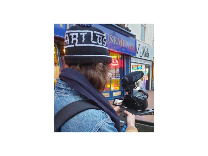 Caroline photographed from behind, filming a street with an amusement centre a nail parlour on it.