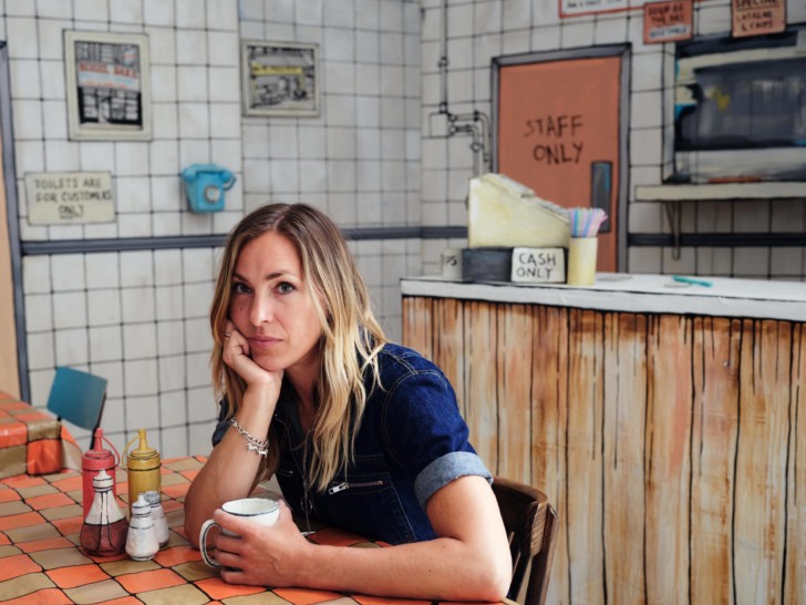 Photograph of a woman at a cafe table. She is wearing a denim shirt and has long blonde hair.