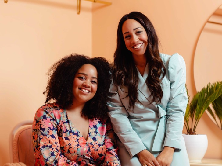 Two women sit together on a sofa, they are smiling and looking at the camera.