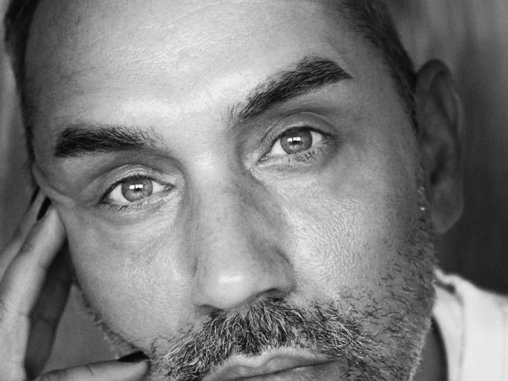 Paul Harleet looks up to the camera in this Black and White headshot, he wears a white t shirt and is resting his head on his hand.