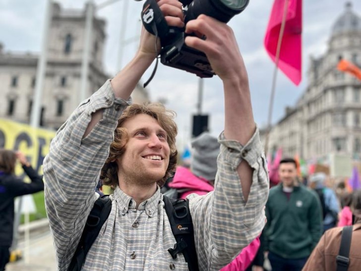 Rich Felgate is photographed filming at a march through a city, he is smiling and wearing a checkered shirt.