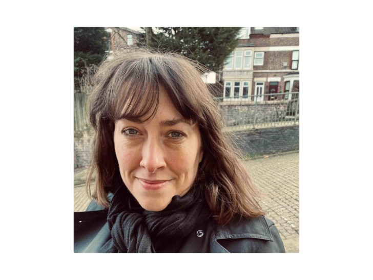 Sarah Bailey stands outside some houses on a cobbled street looking directly into the camera and smiling.