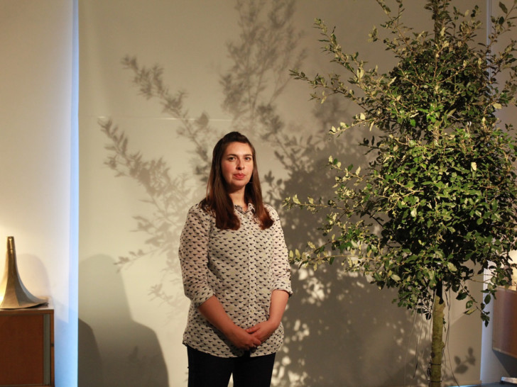 Sheila Ghelani stands in front of an indoor tree in a room with warm lighting. They have long straight brunette hair and hear a polka dot blouse.