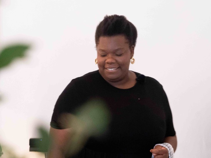 Xhi Ndubisi stood in front of a plant holding a tea towel, smiling at the ground.