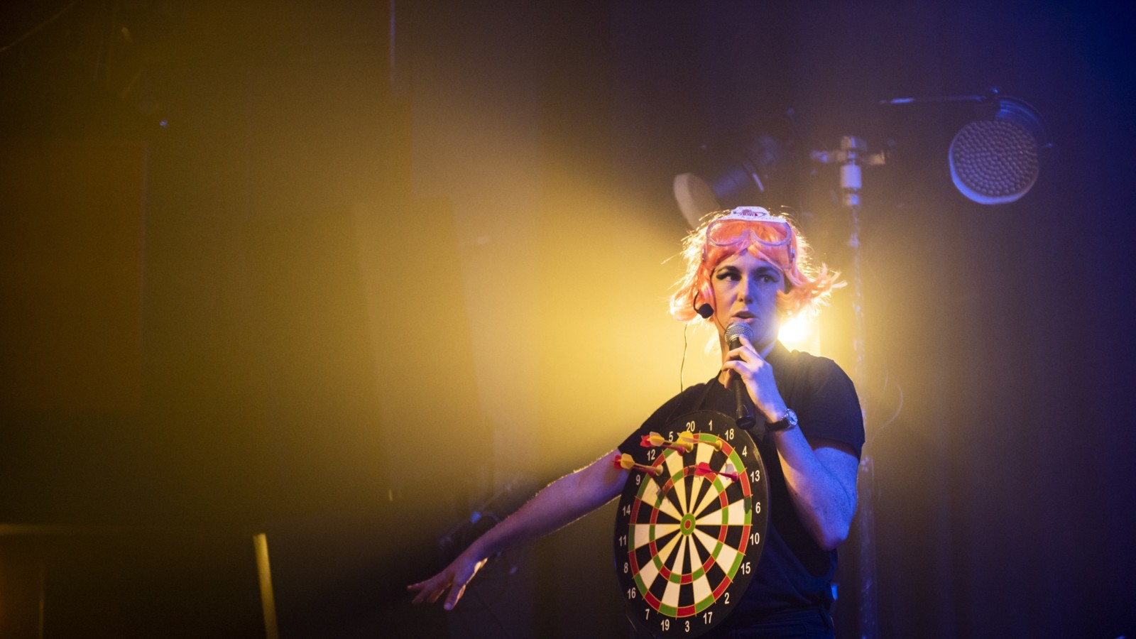 Amy Pennington is dressed in black and is wearing a bright pink wig. They have a dart board hung around their neck so that the target rests on their chest. They are on stage performing, holding a microphone.