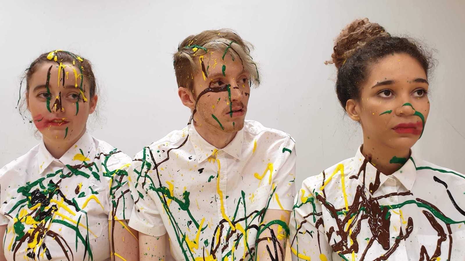 The three members of the 'Year of the Rat Collective' sit next to each other in a line. The are all wearing white shirts and are splattered with green, yellow and back paint