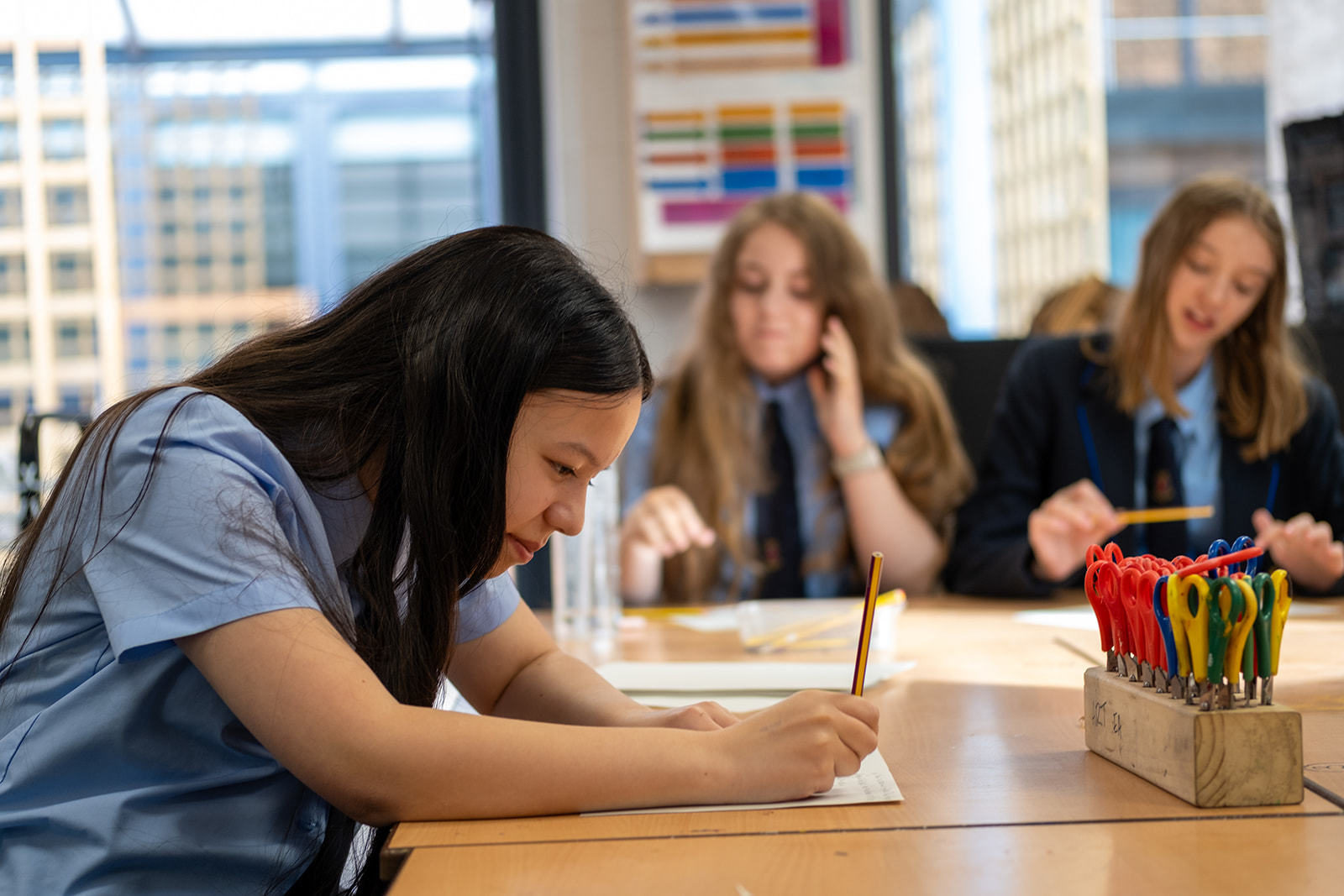 A girl with long dark hair is photographed smiling whilst writing something down in pencil. There are two other students in the background sat at the table.