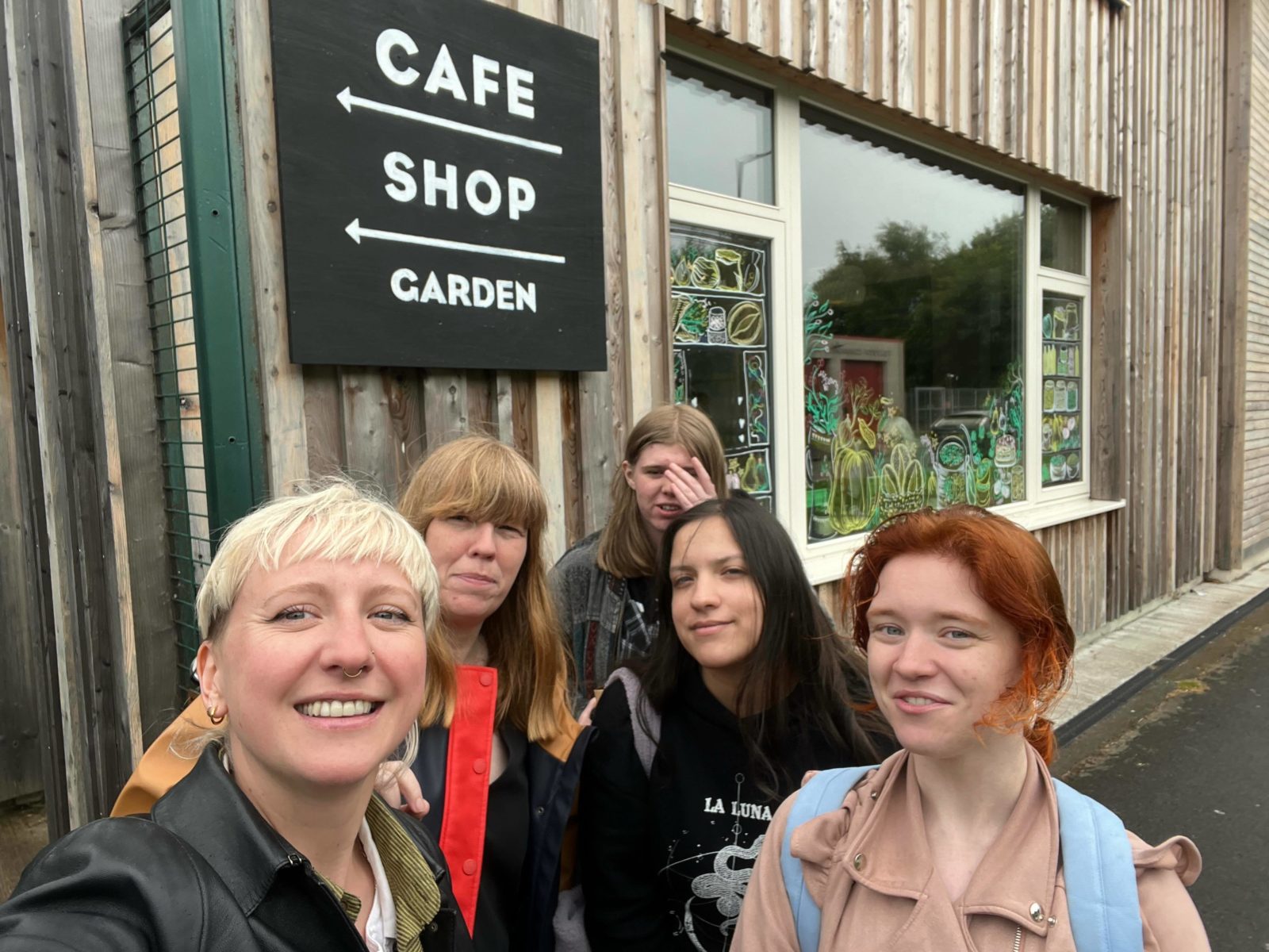 Heart of Glass Producer Emma, Artist Fox Irving and 3 members of the Youth Advisory Group smile for the camera in a selfie. Behind them is a sign with arrows reading 'Cafe, shop, garden.'