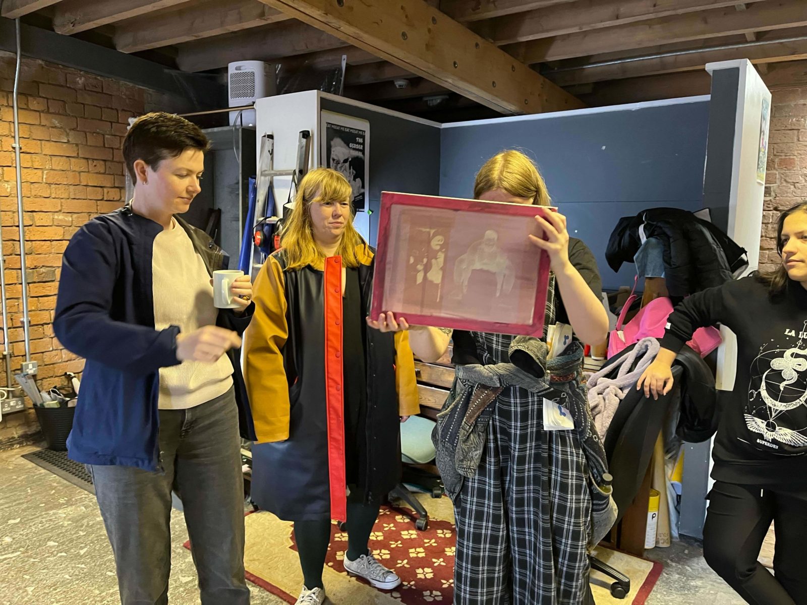 4 people stand in a print room, one is holding up a mesh screen with designs on it and looking at them closely. Another stands with a mug in one hand and others also look at the screen from afar.