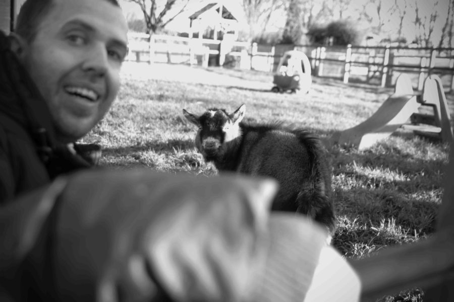 A black and white image of person with short dark hair smiles towards the camera, behind them in a goat.