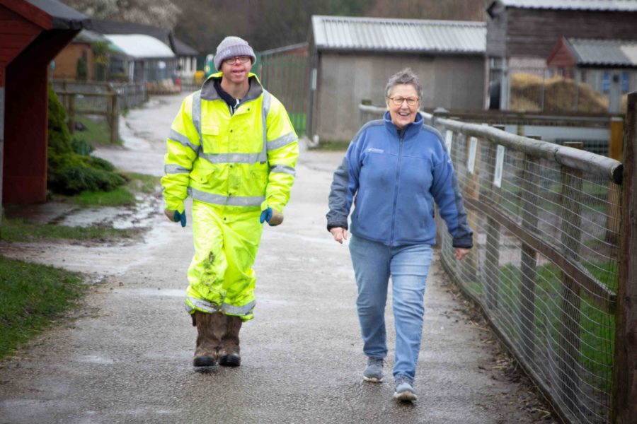 A person in full yellow high vis overalls and another in jeans and a fleece walk along a tarmac path in the rain, smiling