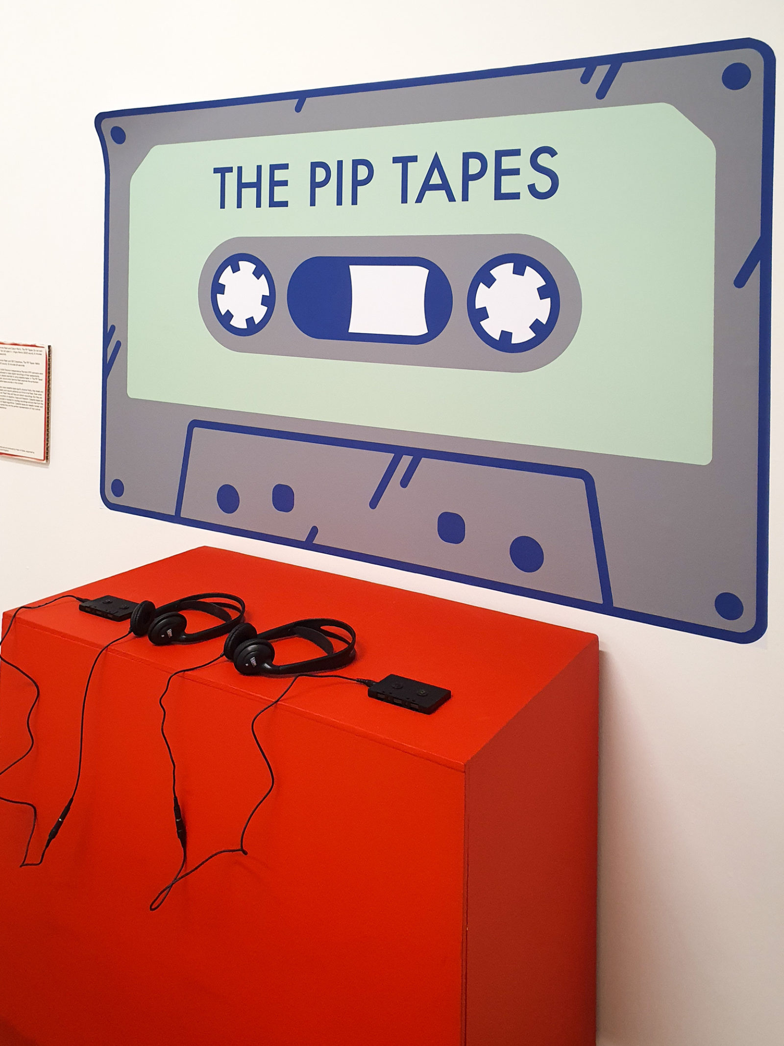 A photo of 'The PIP tapes' set up at the People's History Museum in Manchester. There is a red box with 2 sets of headphones plugged into 2 tapes, and a large tape printed on the wall with the words 'The PIP Tapes' on it.