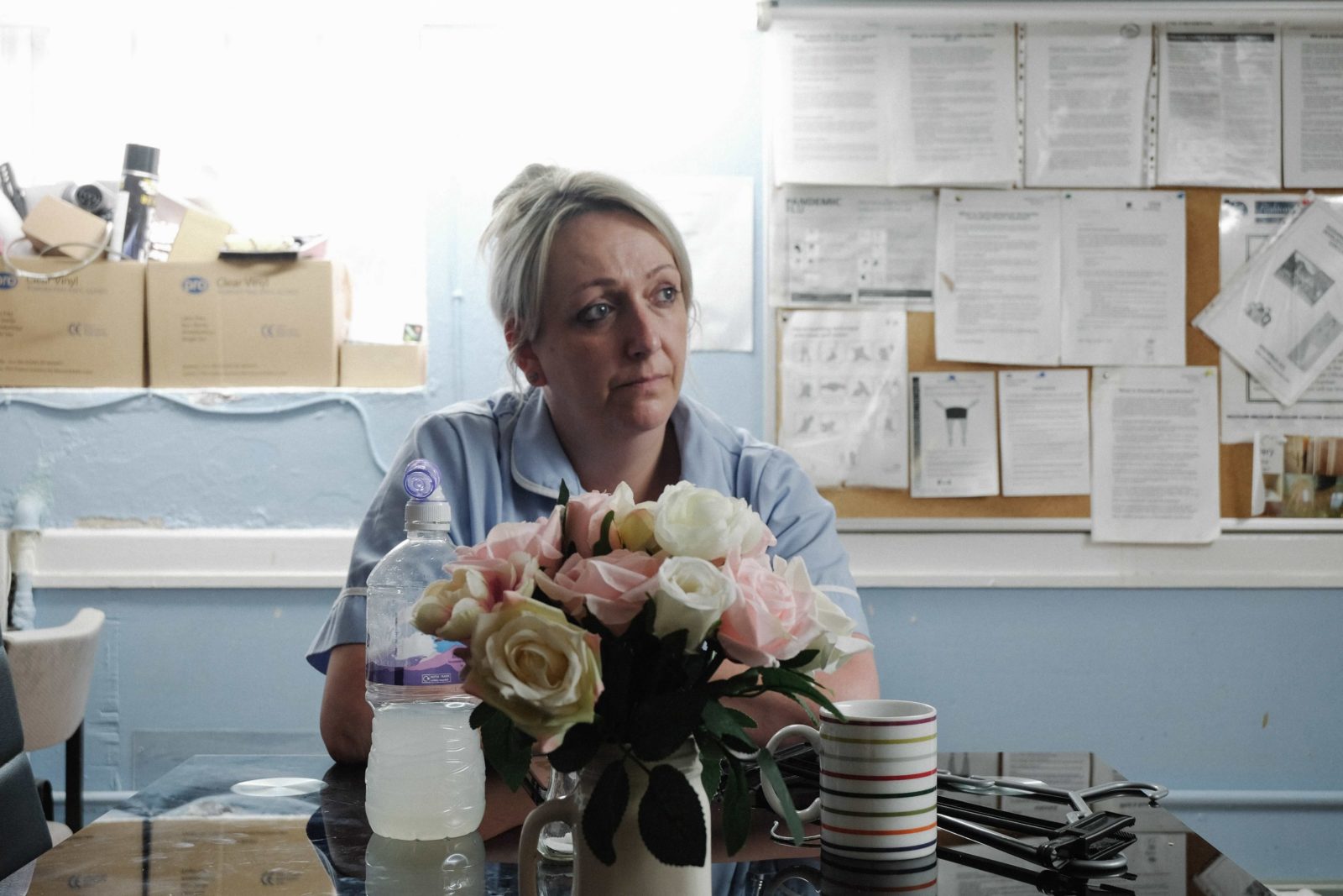 A nurse with blonde hair sits at a desk in a surgery in front of a bunch of flowers. She looks into the distance, looking worried and sad.