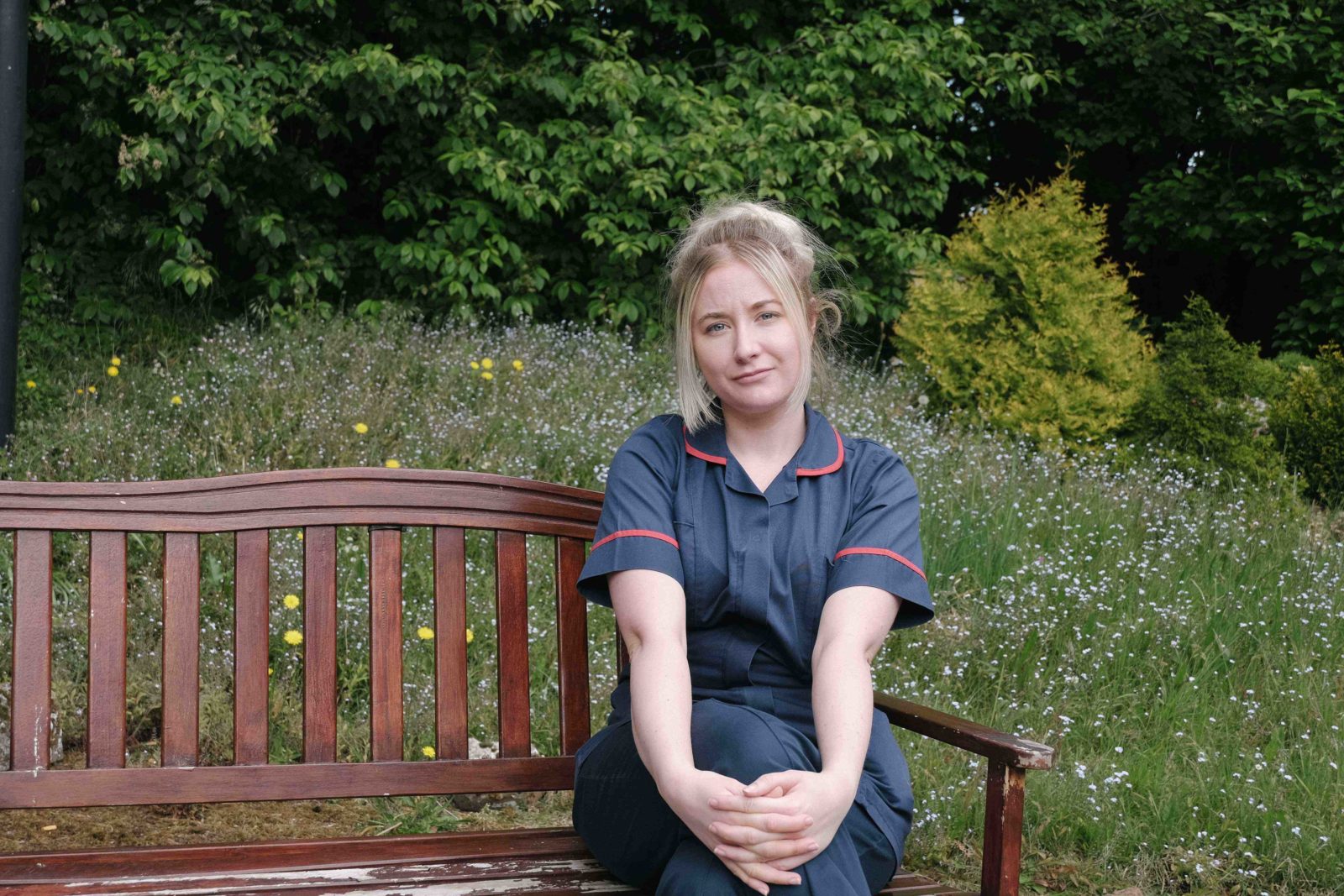 A young blonde woman with her hair tied back half smiles at the camera whilst sat on a wooden park bench, hands clasped around her left knee. She is wearing a nurses or carers uniform.