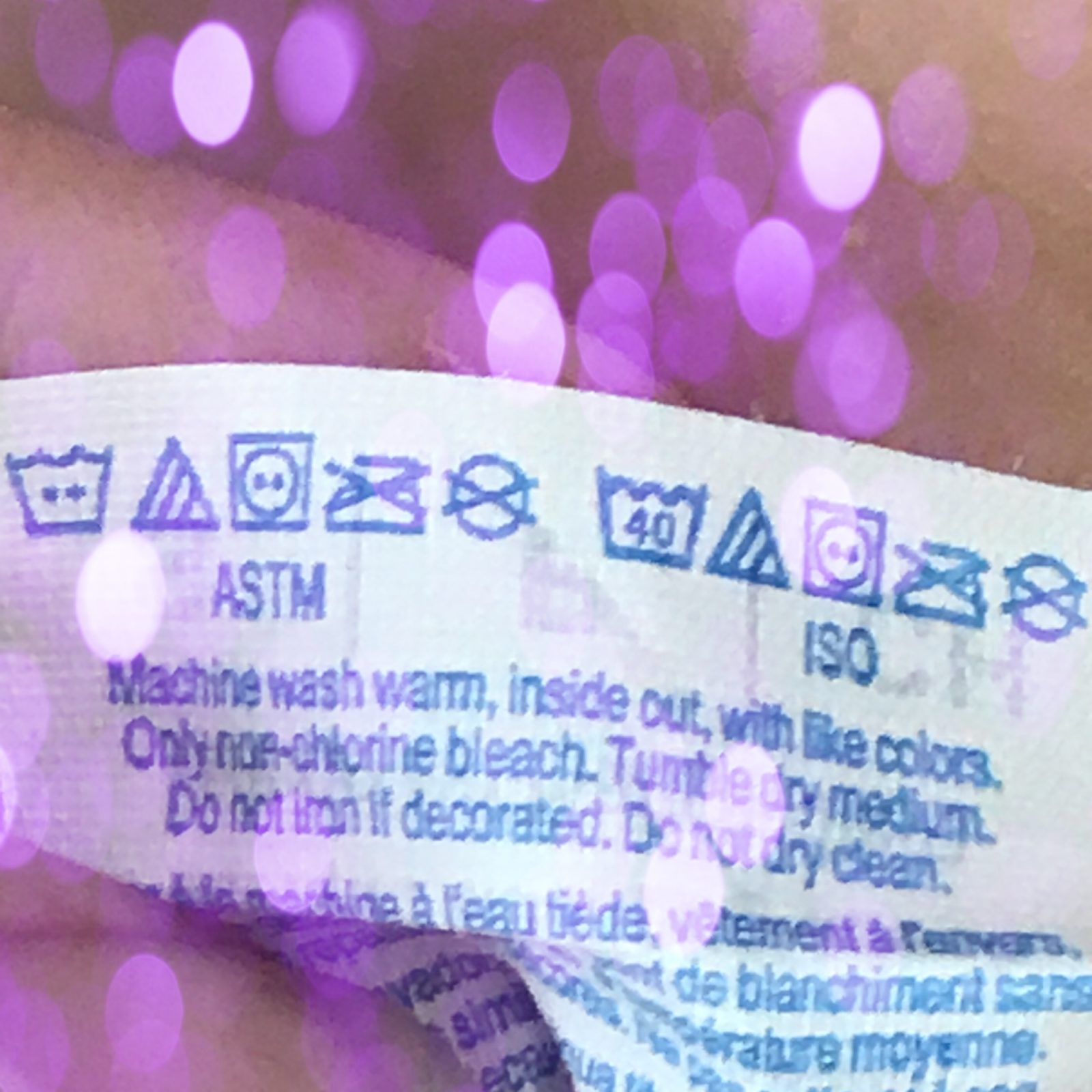 A close up photograph of a clothes label with washing instructions. It is slightly abstracted by pink glitter which is overlayed on top of it.