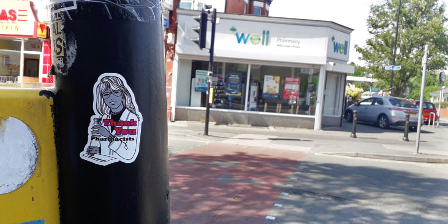 A sticker with an illustration of a pharmacist holding a bottle of tablets that reads 'Thank you Pharmacists' is photographed on a lamppost in front of a pharmacy.