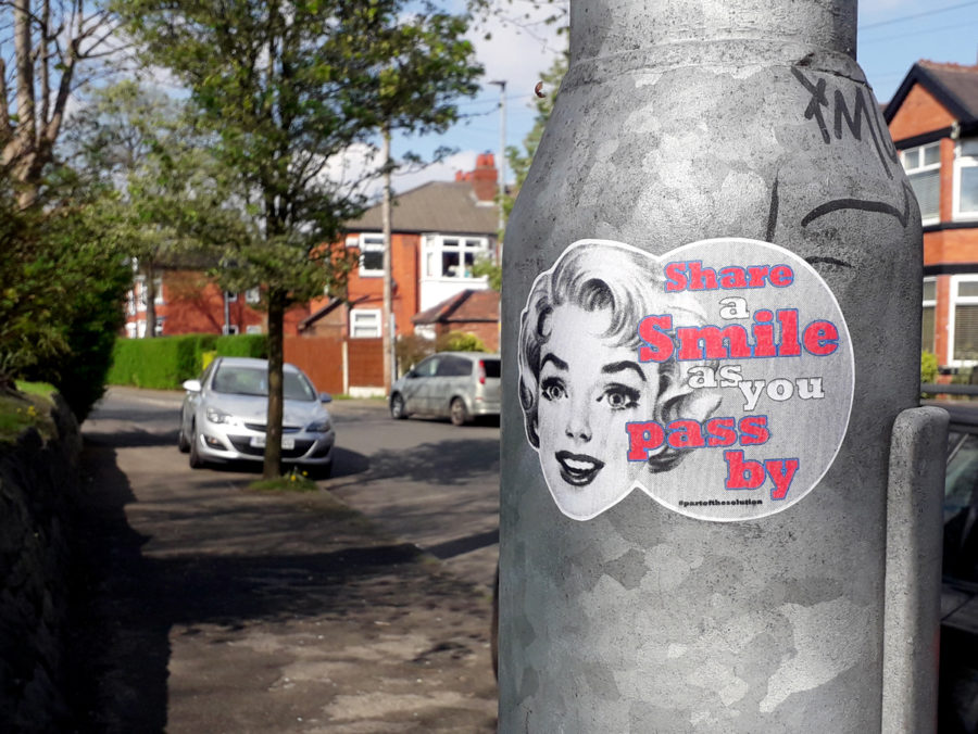 Photo of a sticker on a silver lamppost in English suburbs on a sunny day, the sticker has a black and white photograph of a blonde woman who looks as though she is from the 60's smiling, text next to her face reads; 'Share a smile as you pass by'.