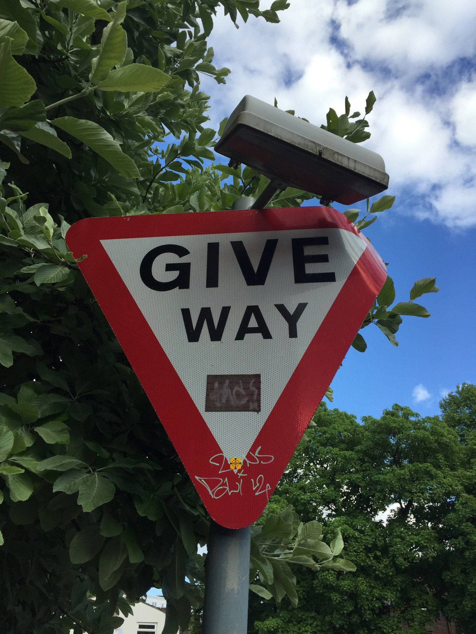 Photograph of a Give Way sign that has been bent backwards at the corner. There is a small sticker of a target at the bottom.