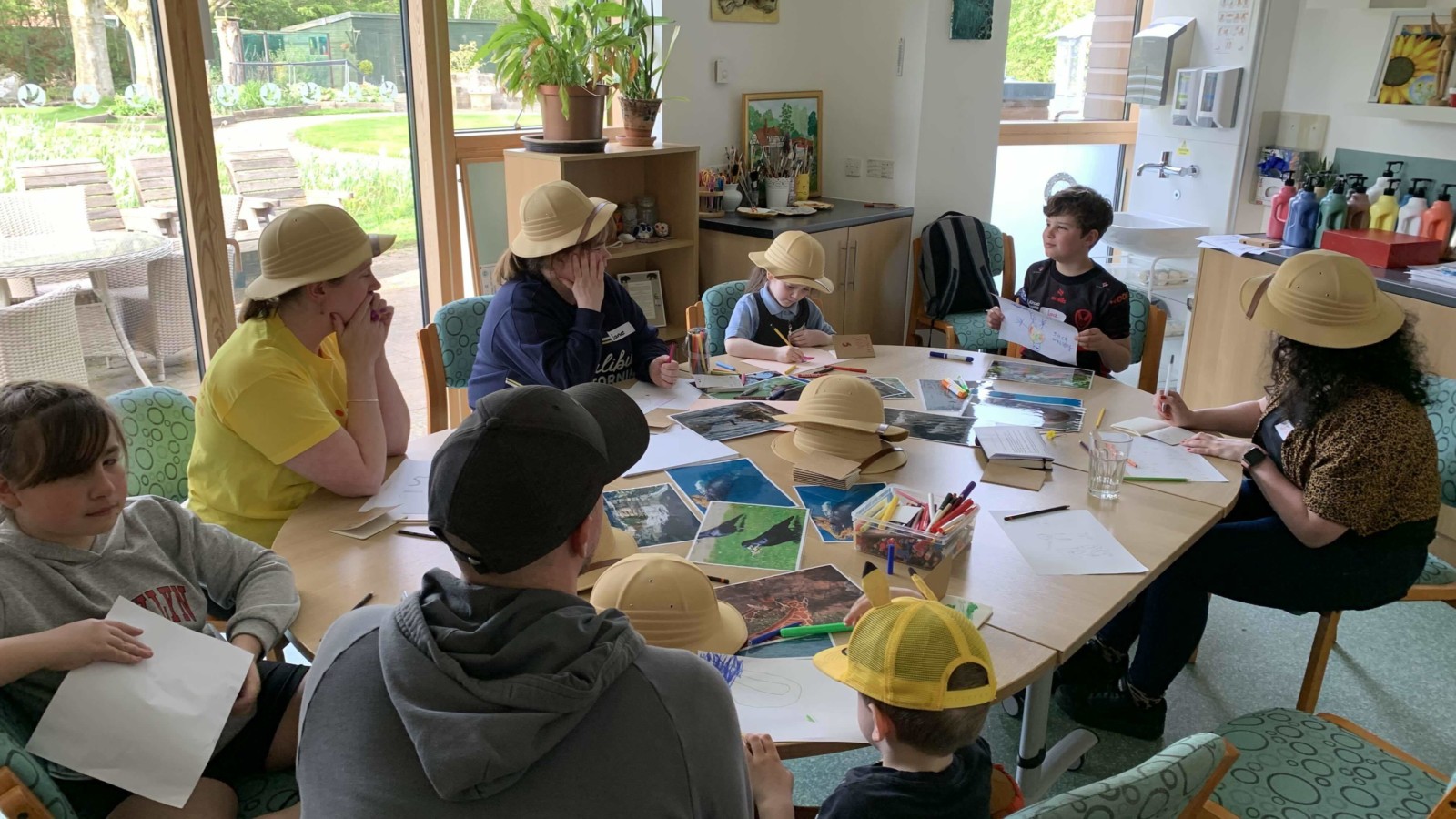 A group of children and adults sit around a table covered in paper and pens. Everyone is wearing a safari hat