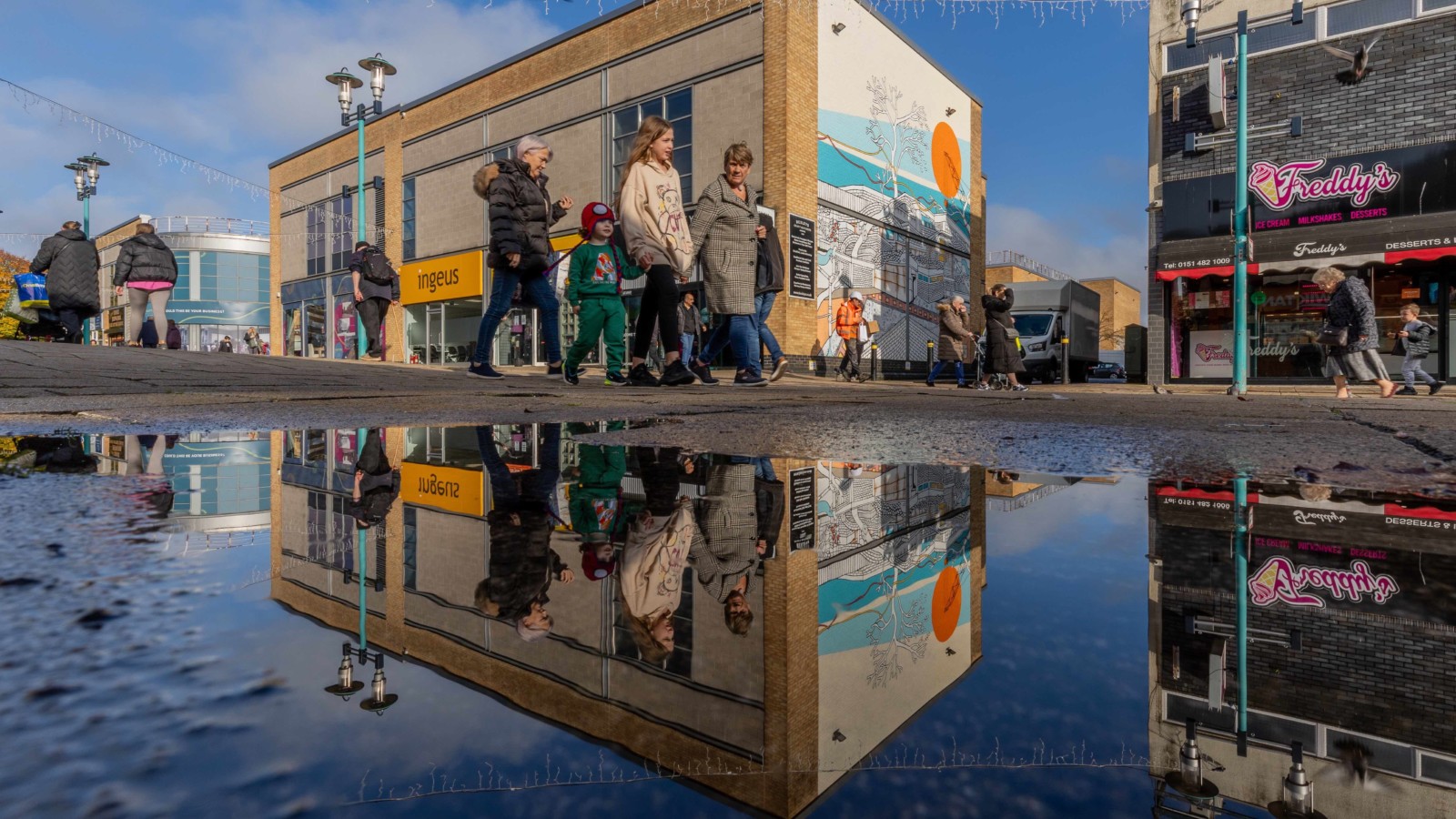 a family walks past the mural on Huyton High Street, their reflections are shown in a puddle below them, and the mural's reflection is behind them.