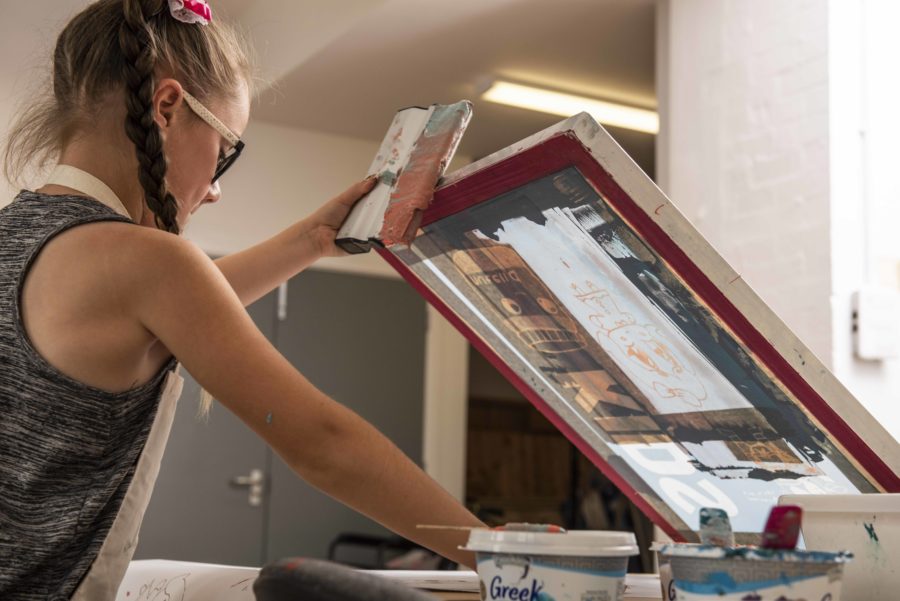 A young person with plaited hair holds a screen printing screen aloft and is surrounded by pots of paint.