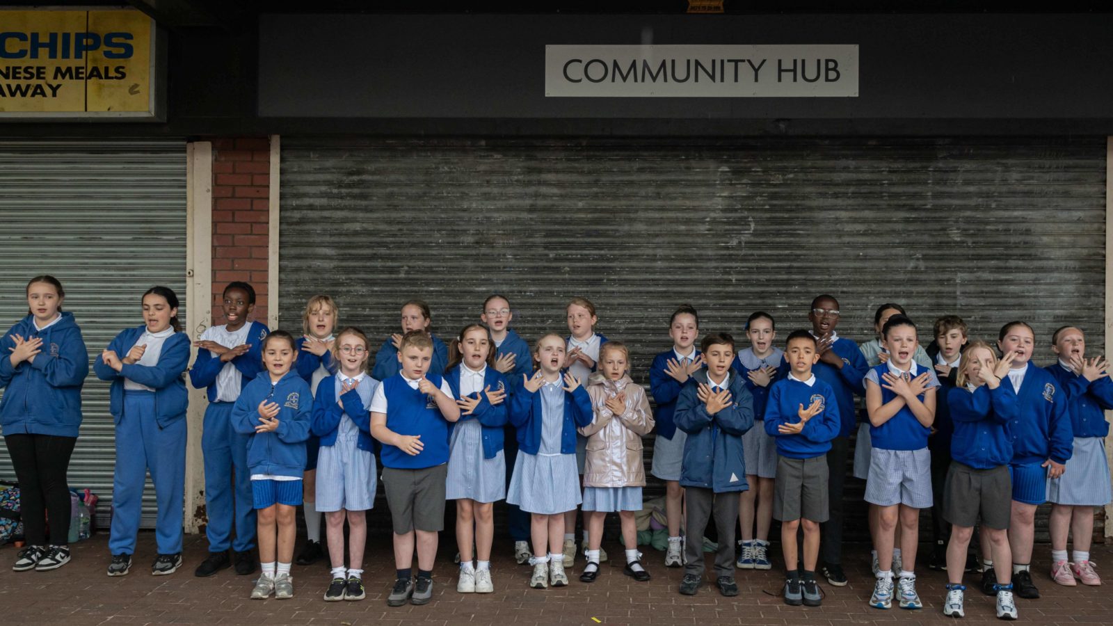 Schoolchildren are in front of a sign that reads ‘Community Hub’ singing and dancing.
