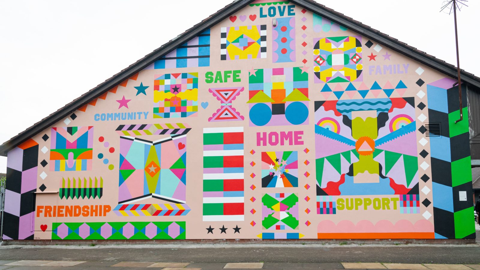 A photograph of the mural up front. The words ‘Friendship’, ‘Family’, ‘Home’, ‘Support’, ‘Love’, and ‘Safe’ can be read amongst abstract, geometric patterns in the colours, lilac, orange, red, blue, green, pink and yellow.
