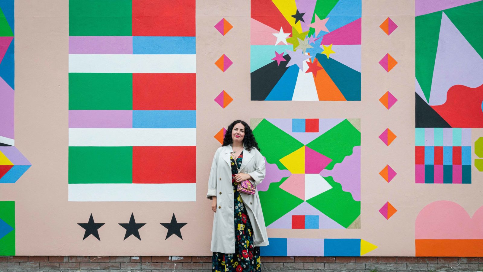 Cherie smiles at the camera in front of the mural. She has long, black curly hair and wears a floral dress with a raincoat over it and red lipstick.