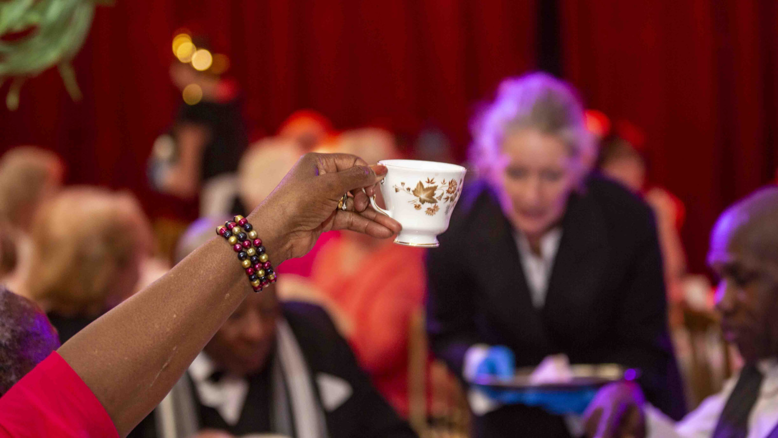 A black person wearing a red shirt and shiny bracelet holds up a posh tea cup at a posh afternoon tea event