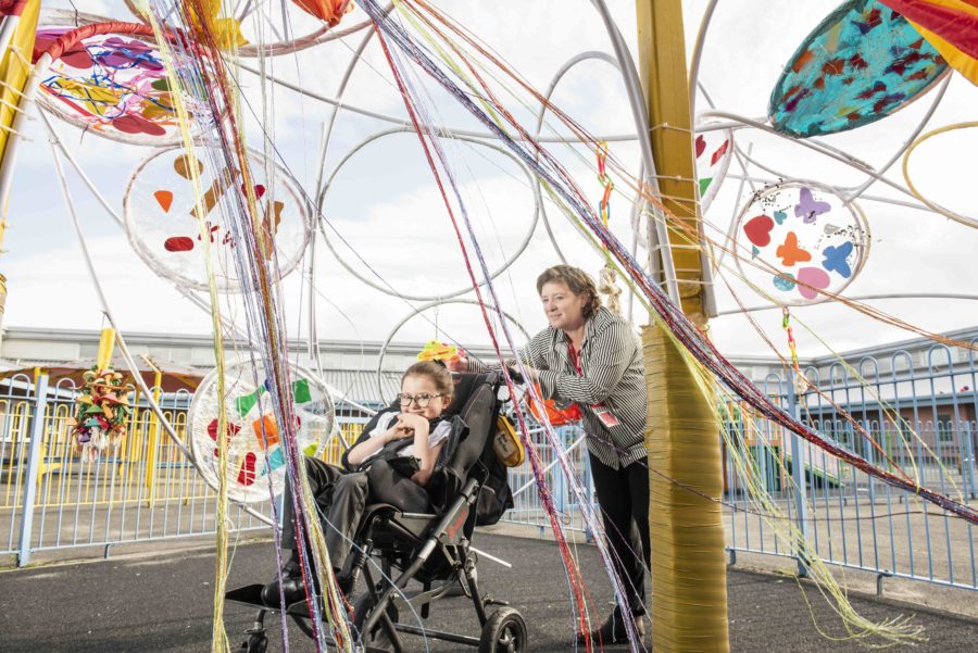 A woman in a stripy shirt is pushing a small girl in a wheelchair underneath a brightly coloured artwork made of streamers and hula hoops in a playground. The woman is smiling and the young girl is grinning.