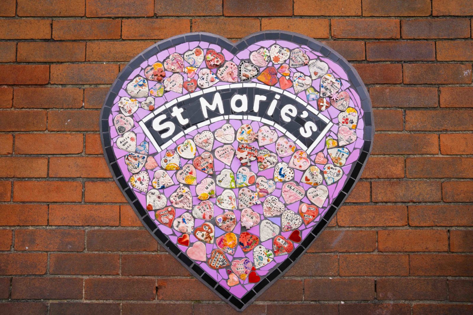 A heart shaped mosaic is cemented to a red brick wall. It has a lilac background and is filled with heart shaped tiles with names, words and decorations on them. In the middle it reads ‘St Marie’s’ in an arc shape with a black background and white letters.