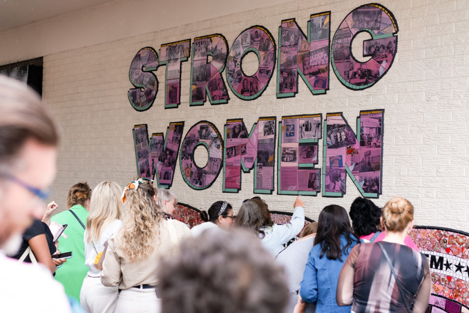 A crowd of women crowd around the mural, pointing at the strong Women of Knowsley Mural and taking photos.