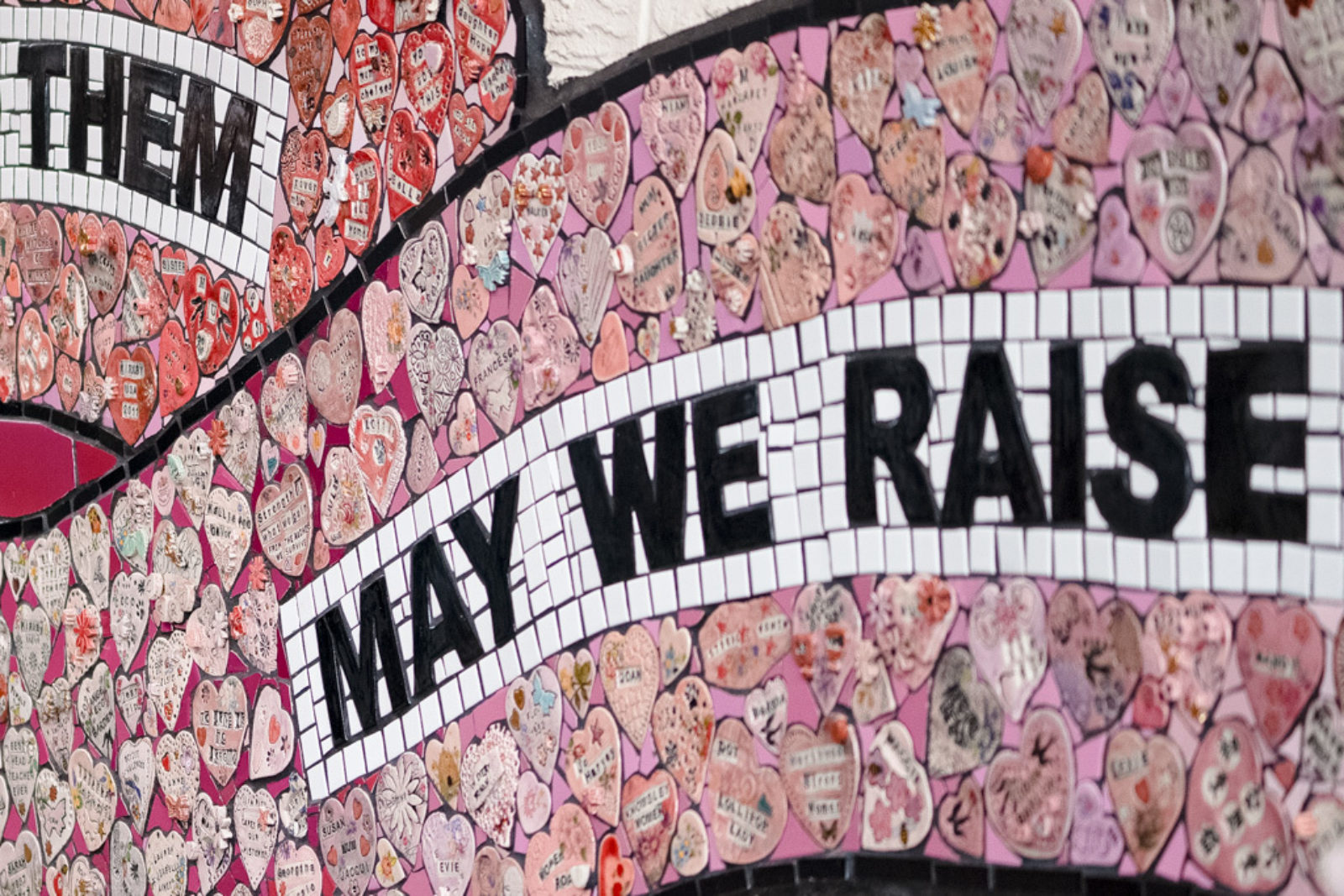 The mural is photographed up close, showing two banner shapes that read ‘May we raise’ surrounded by clay hearts.
