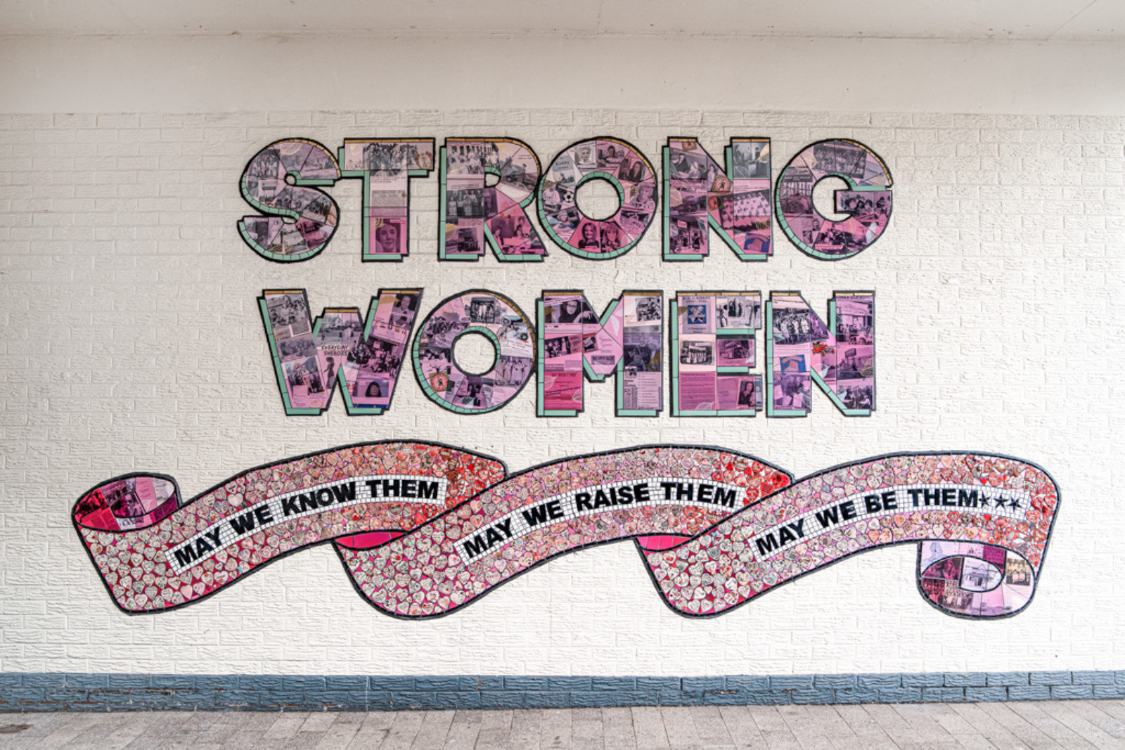 The strong women of knowsley mural is photographed in Kirkby. It is on a white brick wall, and reads ’Strong Women’ In tiled pink capital letters with newspaper clippings and black and white photographs printed on them. Underneath is a scroll reading ‘May We Know them, May we raise them, may we be them’ surrounded by clay hearts made by the community, dedicated to strong women in their lives.