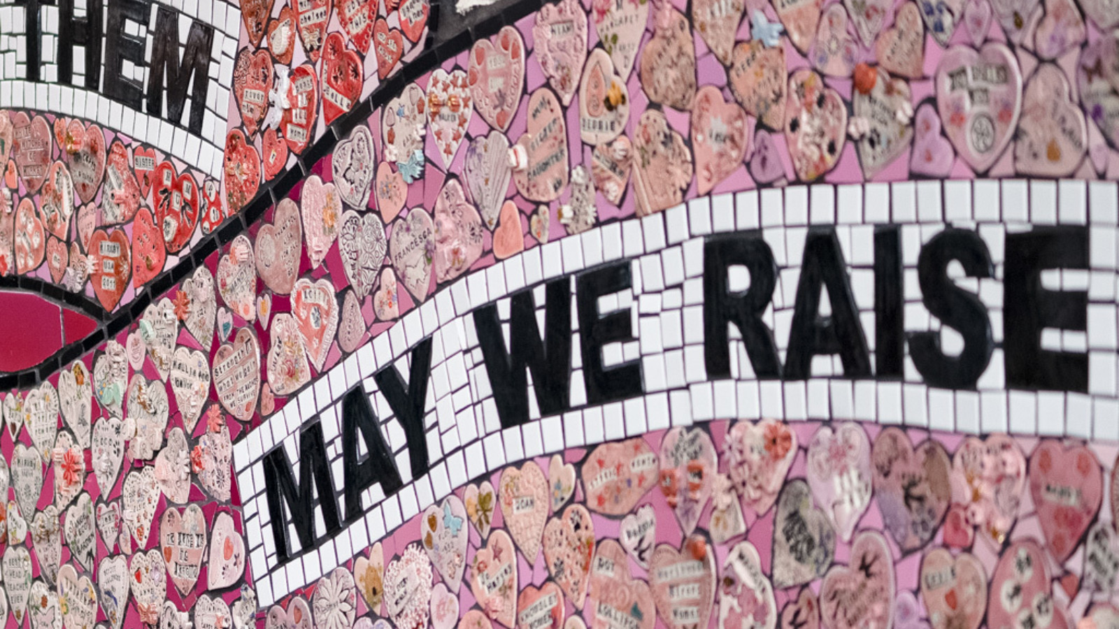 The mural is photographed up close, showing two banner shapes that read ‘May we raise’ surrounded by clay hearts.