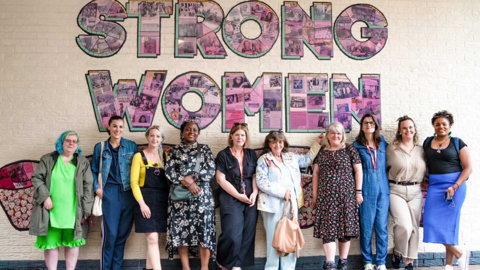 10 women smile for the camera stood in front of the mural.