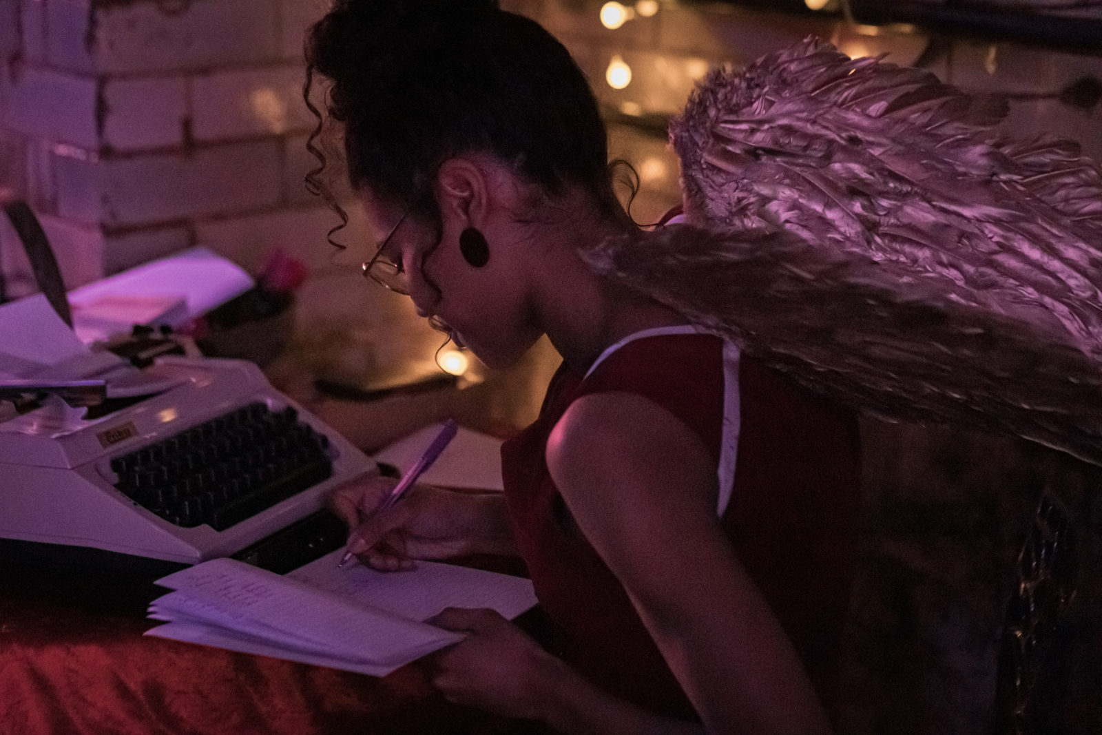 A young woman is sat at a table writing in a notepad. Next to her is a typewriter. She is wearing silvery angel wings.