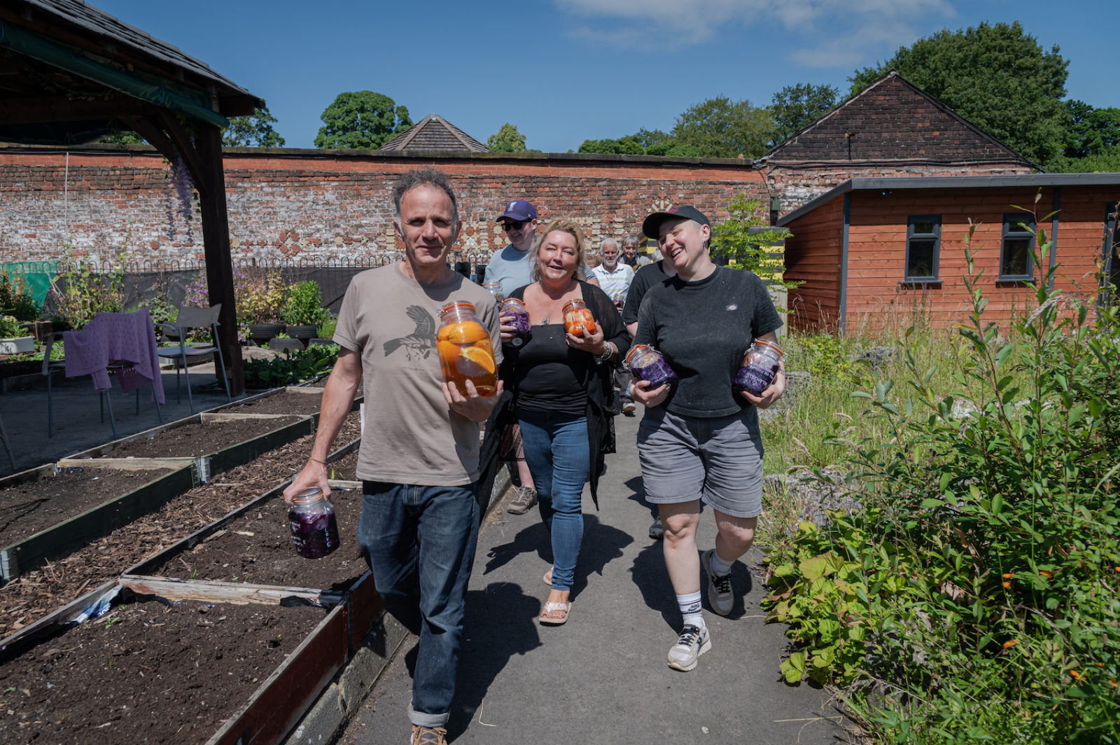 A group of adults walk through a garden, carrying jars of pickles