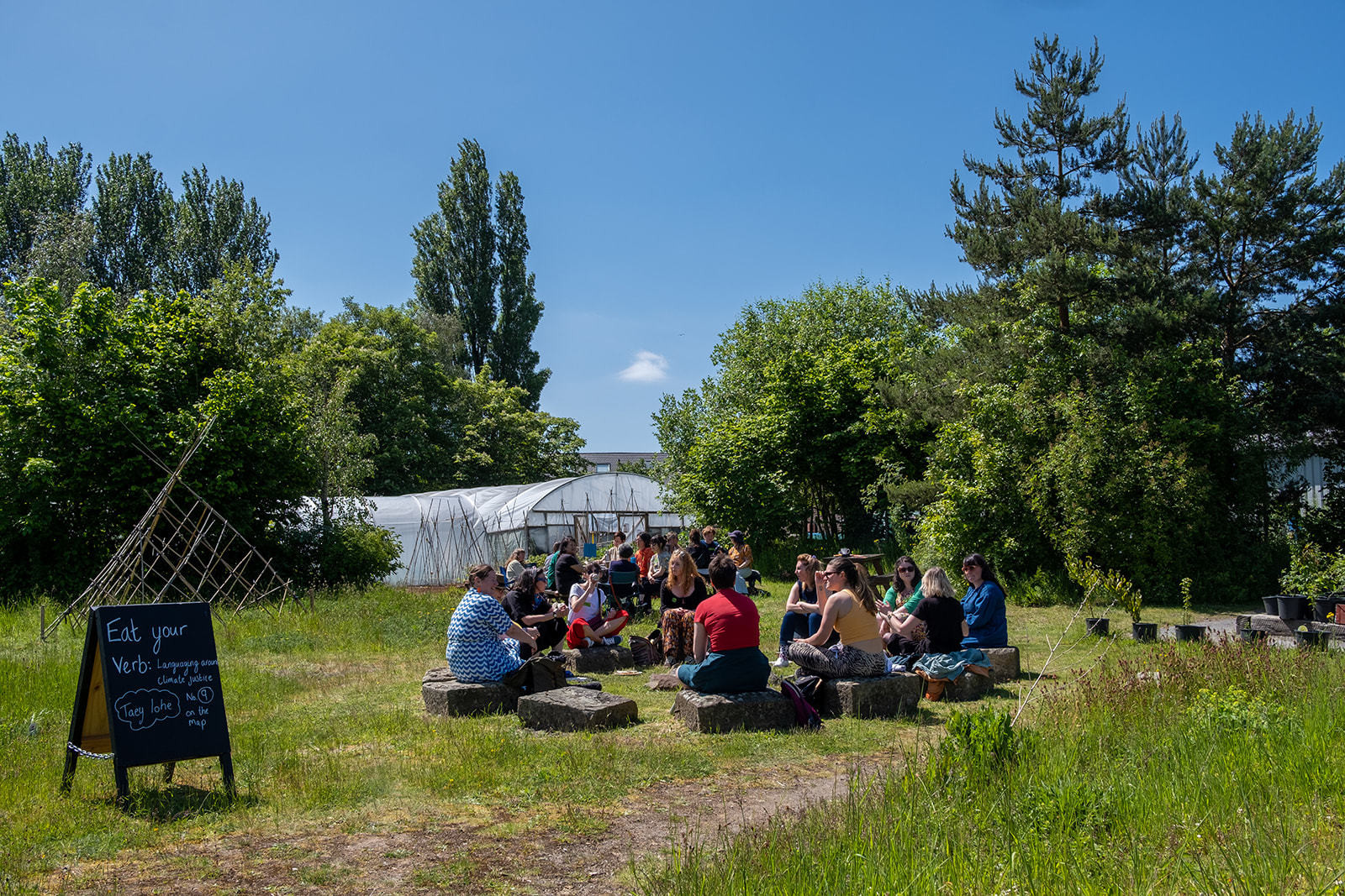 People are sat in the community garden at Incredible Edible Knowsley, it is sunny and there is a sign that reads 'Eat your Verb'