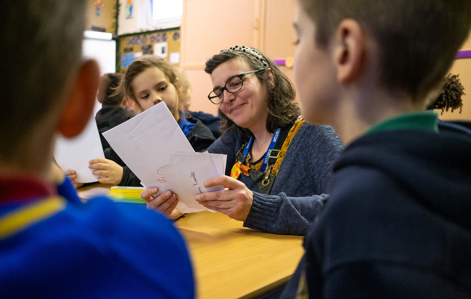 Harriet from interference art holds sheets of paper sat at a table as 3 kids at the table look towards her, she is smiling.