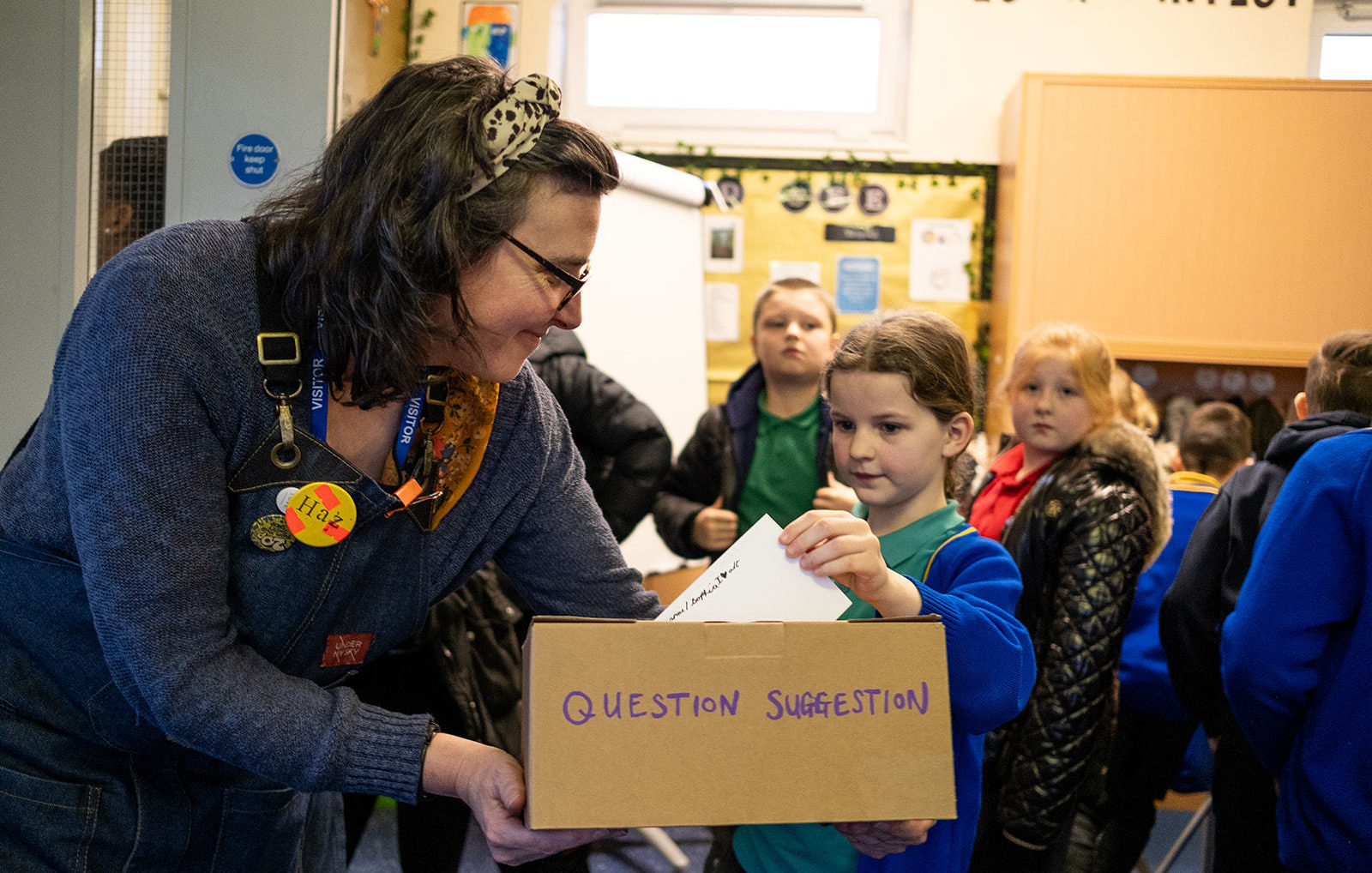 A school child puts a piece of paper into a cardboard box that an adult is holding with the words 'Question Section' written on it in blue marker.