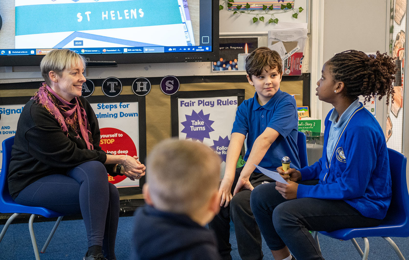 2 school children in blue uniform sit at the front of the classroom talking to a teacher.