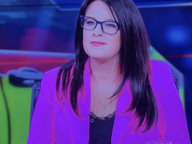 Photograph of a woman wearing a pink blazer and black rimmed glasses, with dark brown hair