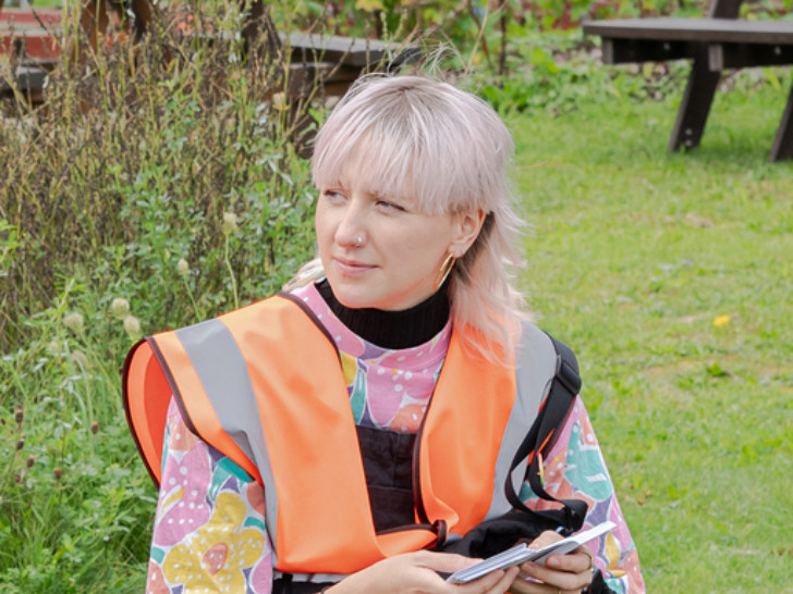 Emma wears a high vis vers with a colourful shirt underneath. They have short, light pink hair in a mullet style with a full fringe.