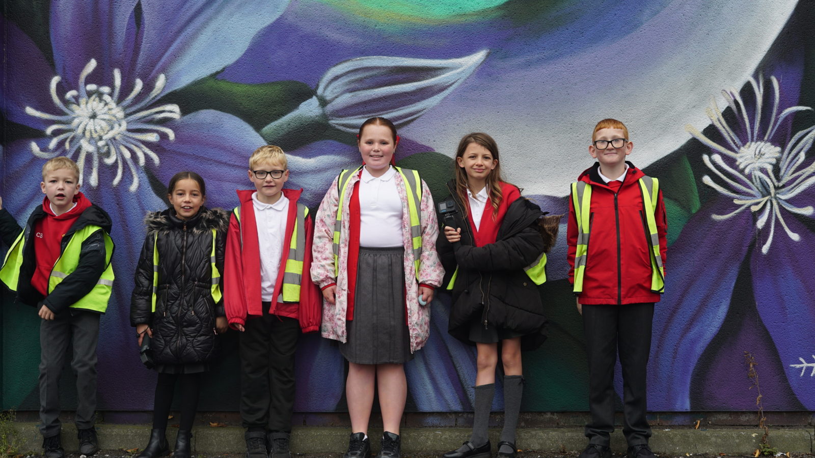 Photograph of a group of children in red school uniforms, standing in front of a colourful mural. They are all smiling.